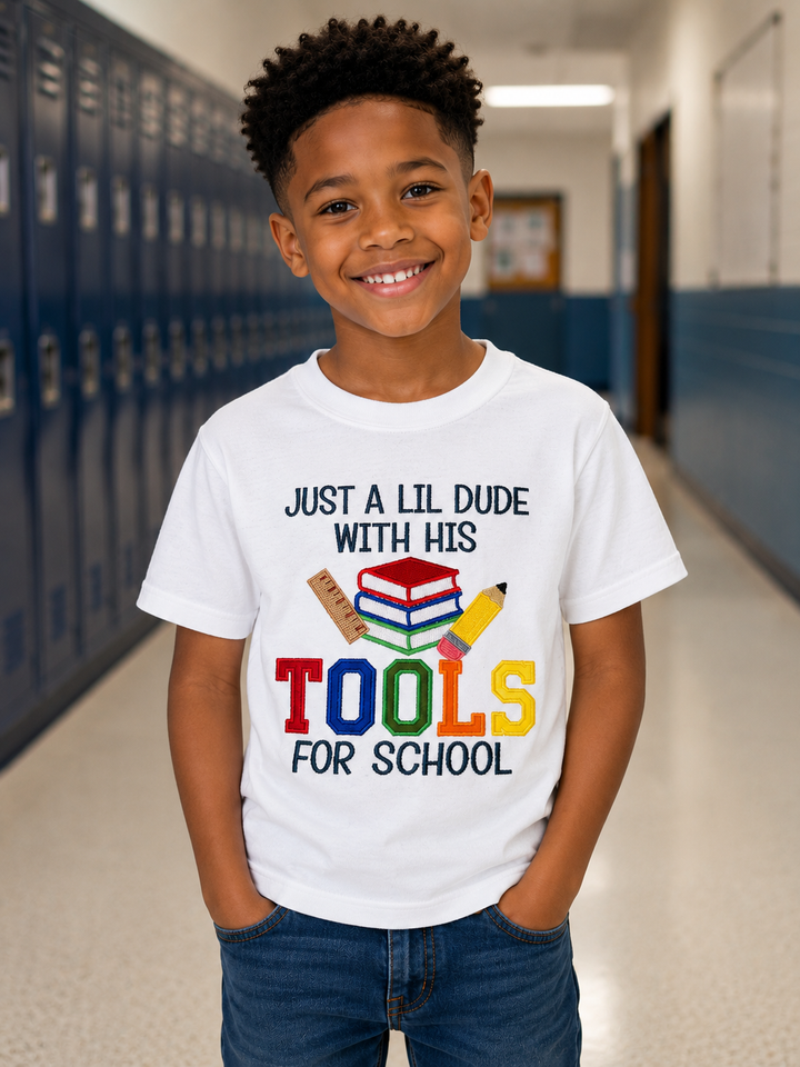 Boy wearing a white embroidered “Just a Lil Dude with His Tools for School” shirt featuring colorful appliqué letters and school supplies for back to school.