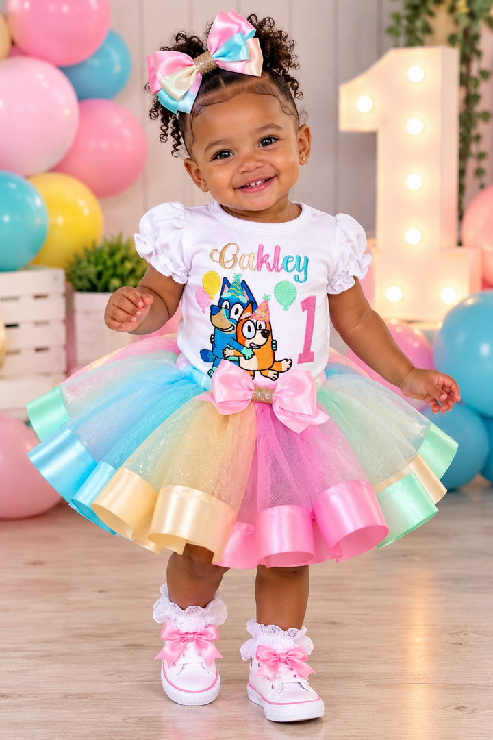 A toddler girl wearing a colorful first birthday costume with a white ruffled top, a pastel tulle tutu skirt in blue, pink, yellow, and green, and a large pink bow. She is smiling in a decorated party setting with balloons and a large number 1 backdrop.
