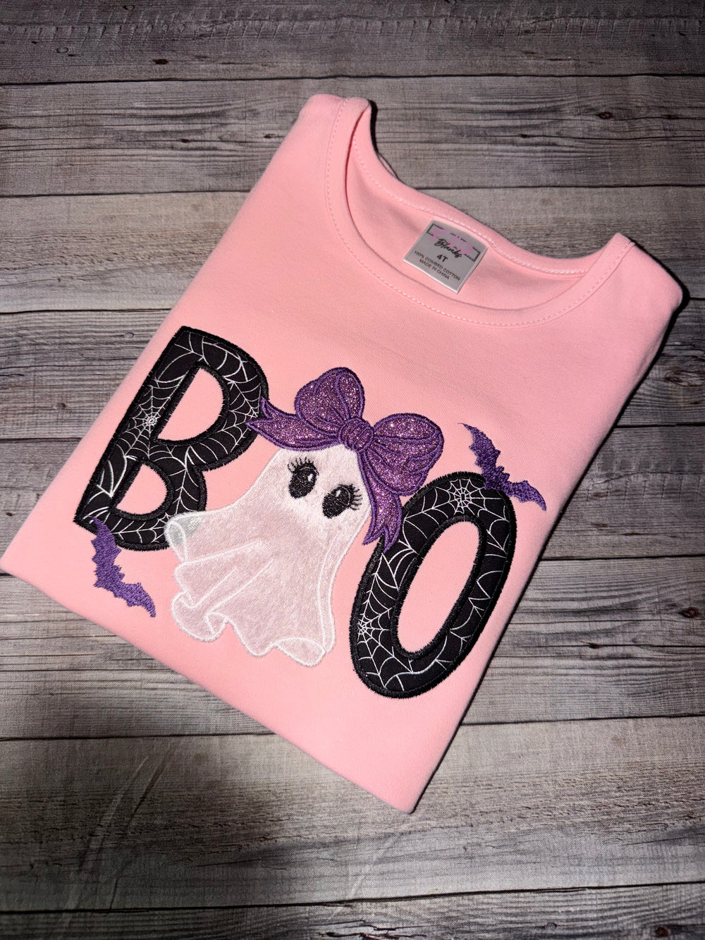 Pink girls Halloween shirt featuring an embroidered ghost with a glitter purple bow, spiderweb Boo appliqué letters, and embroidered bats.