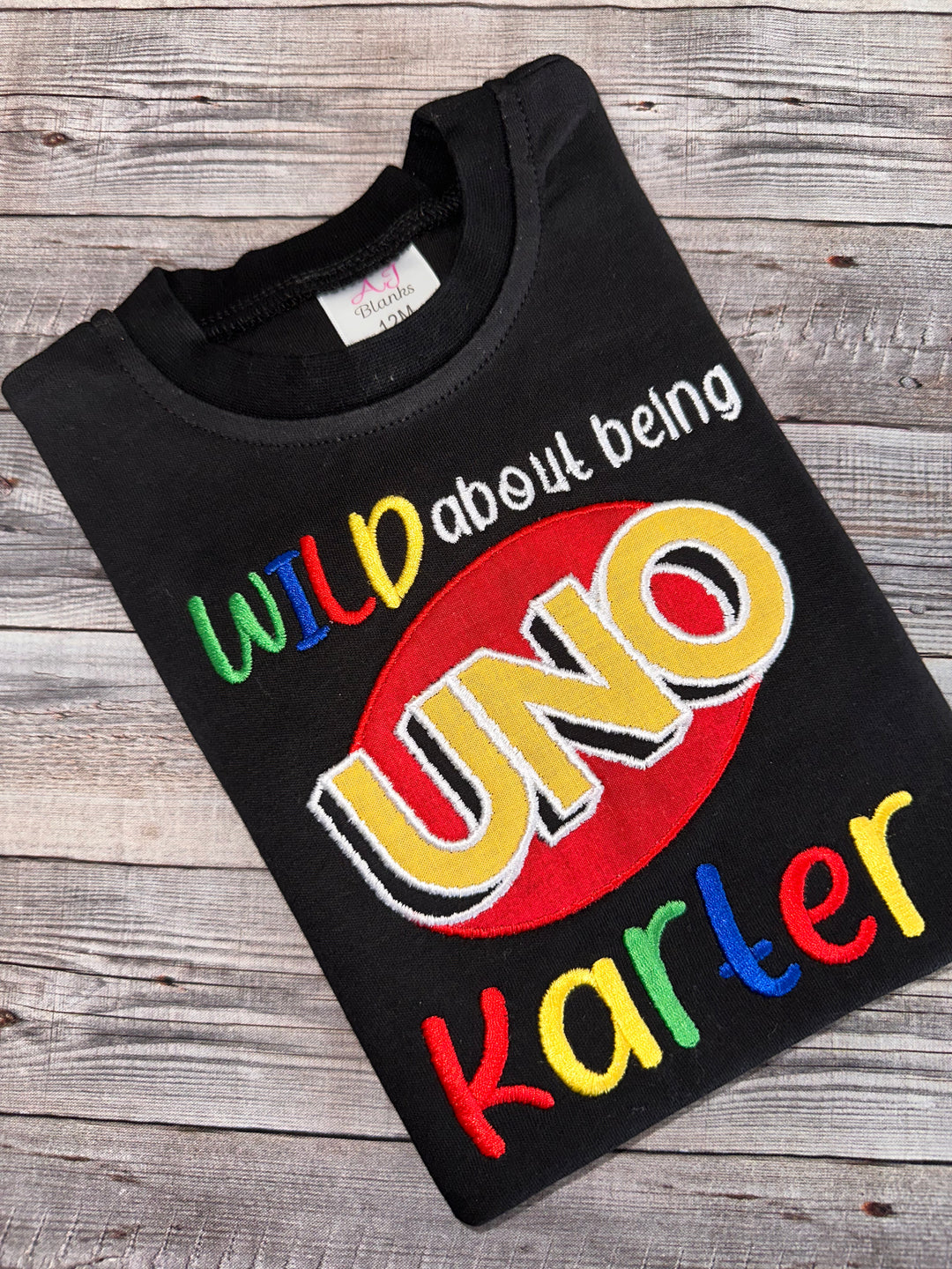 Black personalized Wild About Being UNO first birthday shirt featuring colorful embroidery, UNO logo appliqué, and custom name for a boy’s first birthday celebration.