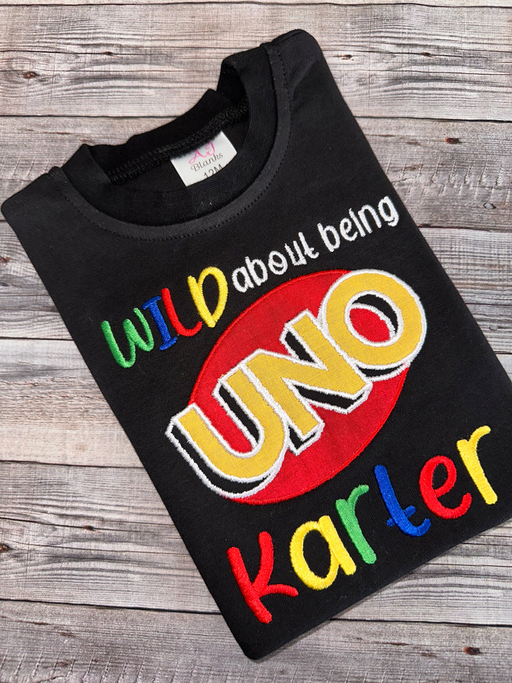 Black personalized Wild About Being UNO first birthday shirt featuring colorful embroidery, UNO logo appliqué, and custom name for a boy’s first birthday celebration.