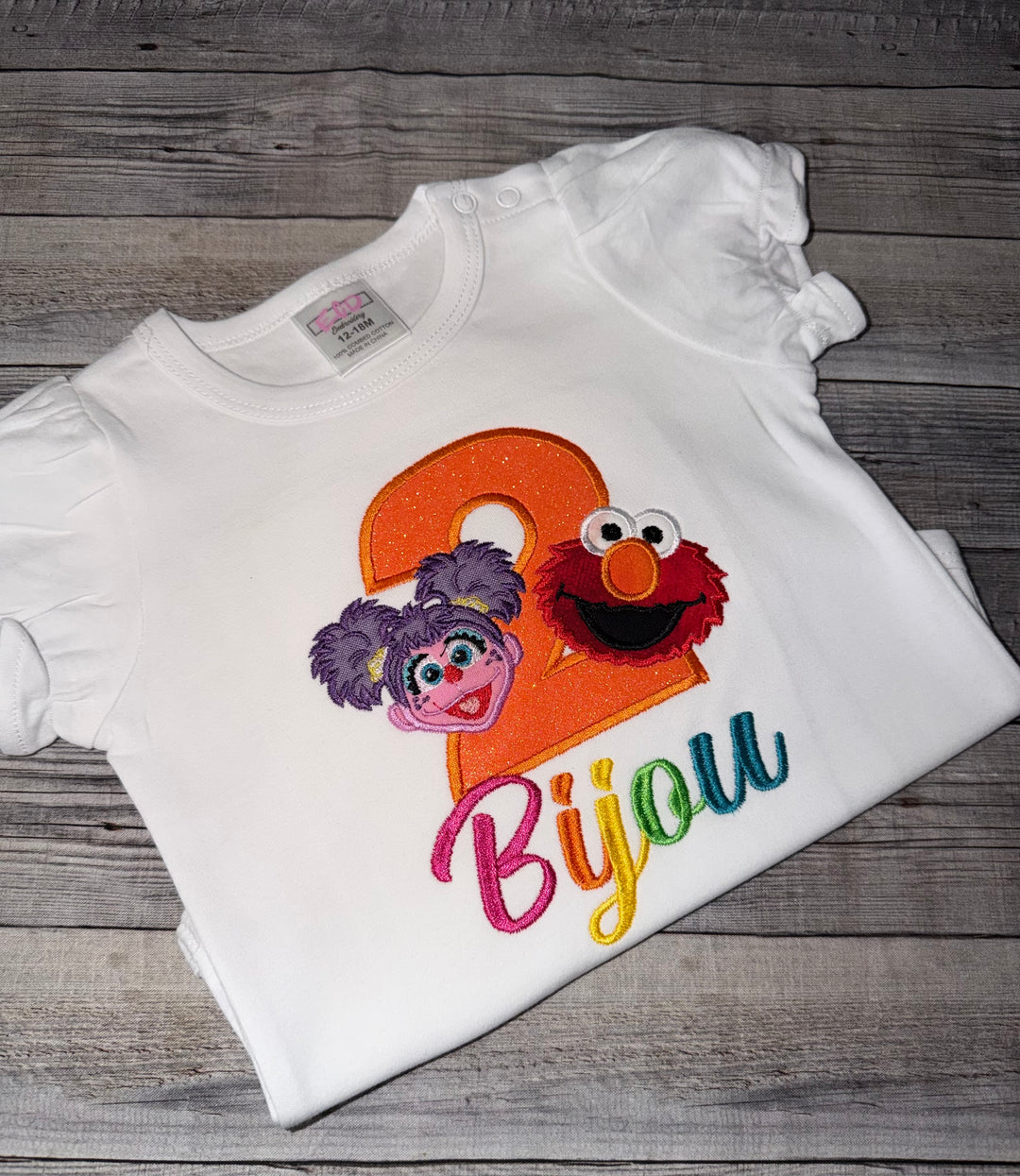 White shirt with Sesame Street-themed design featuring Elmo and Abby Cadabby on a wooden surface.
