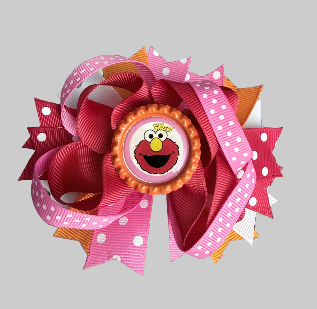 A pink and red hair bow featuring a central circular Elmo button, layered with pink polka dot ribbon, red grosgrain ribbon, and orange and white striped accents, set against a solid gray background.