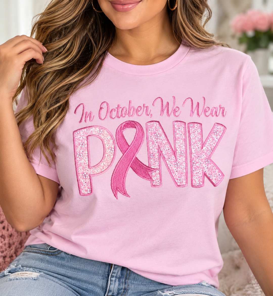 Pink embroidered In October We Wear Pink crewneck sweatshirt featuring floral appliqué lettering and a pink breast cancer awareness ribbon on a rustic wood background.