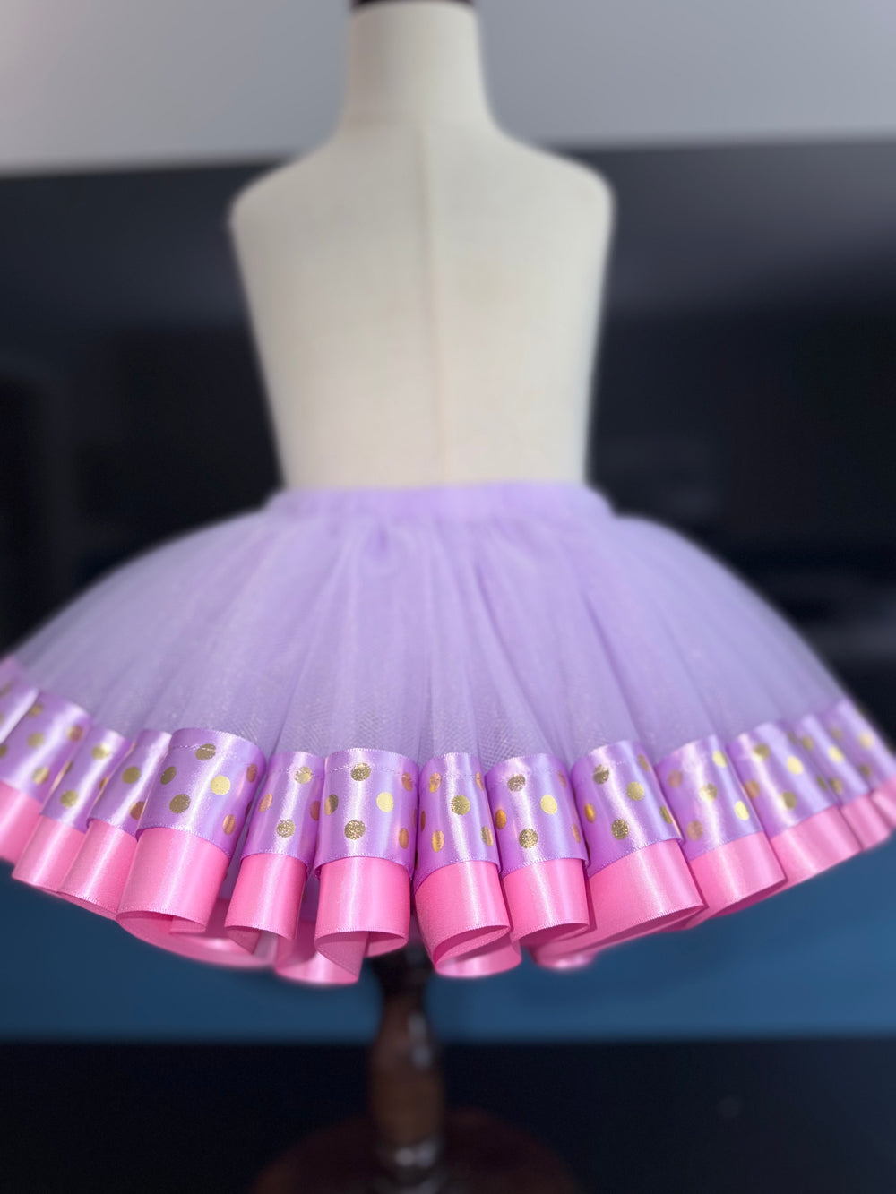 A lavender and pink tutu skirt displayed on a white dress form mannequin. The skirt features a light purple tulle body with a ruffled hem trimmed in pink satin ribbon and decorated with gold polka dots on a purple band.