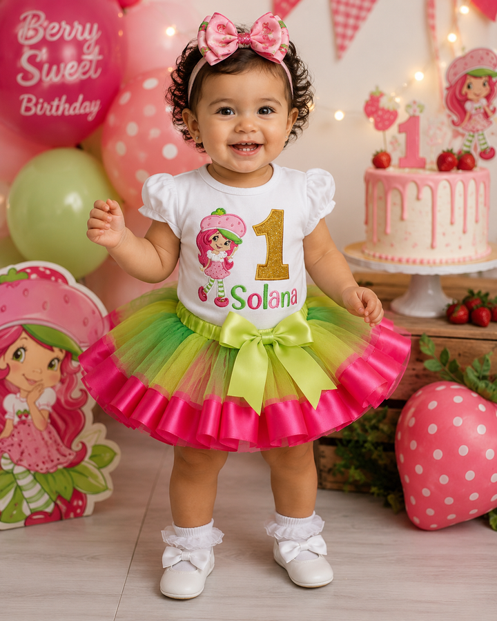 Child in a colorful birthday outfit with a Strawberry Shortcake theme, standing in front of a birthday cake and balloons.