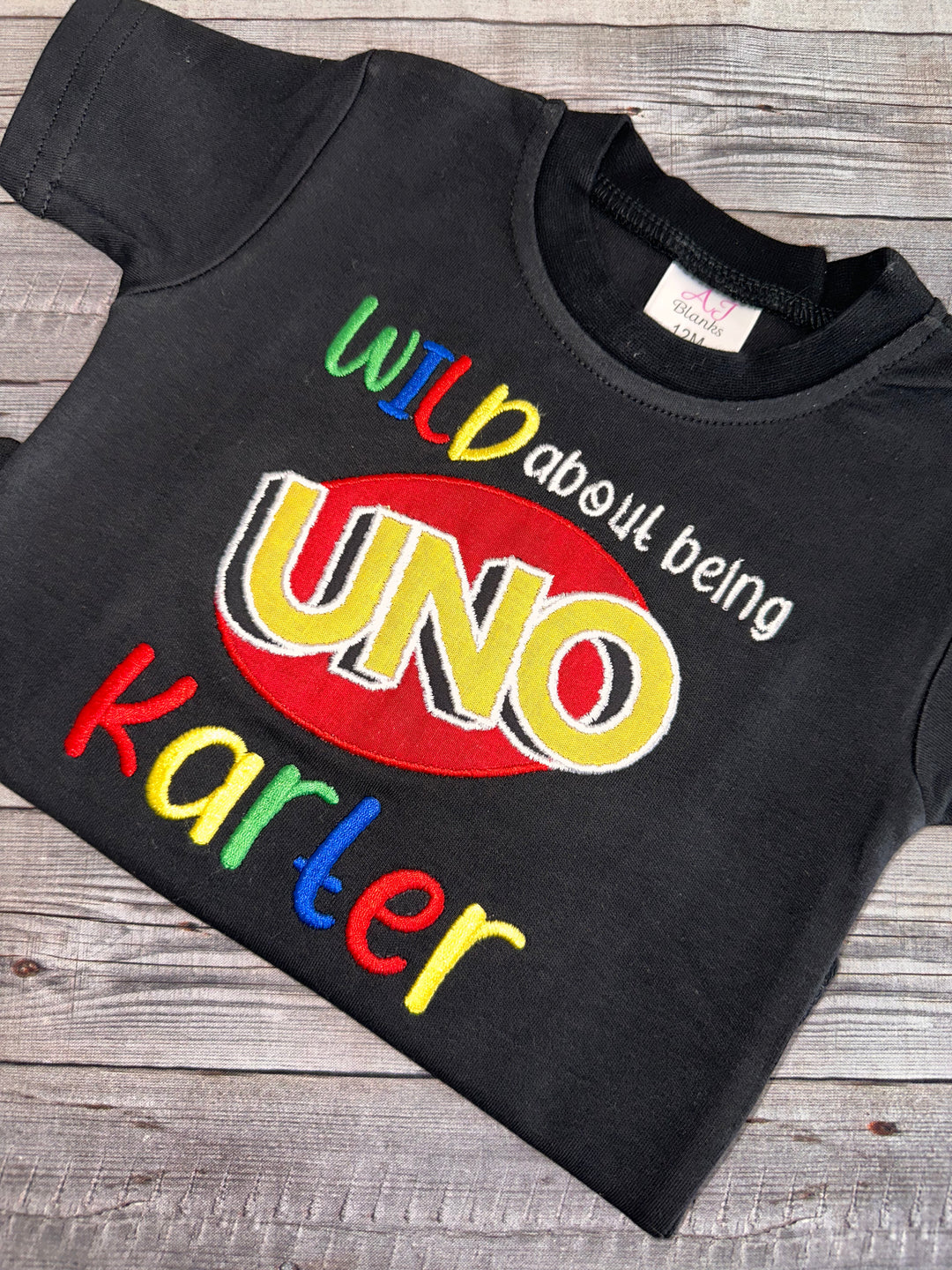 Close-up of a black personalized UNO birthday shirt with colorful embroidered lettering, UNO logo appliqué, and custom child name.”