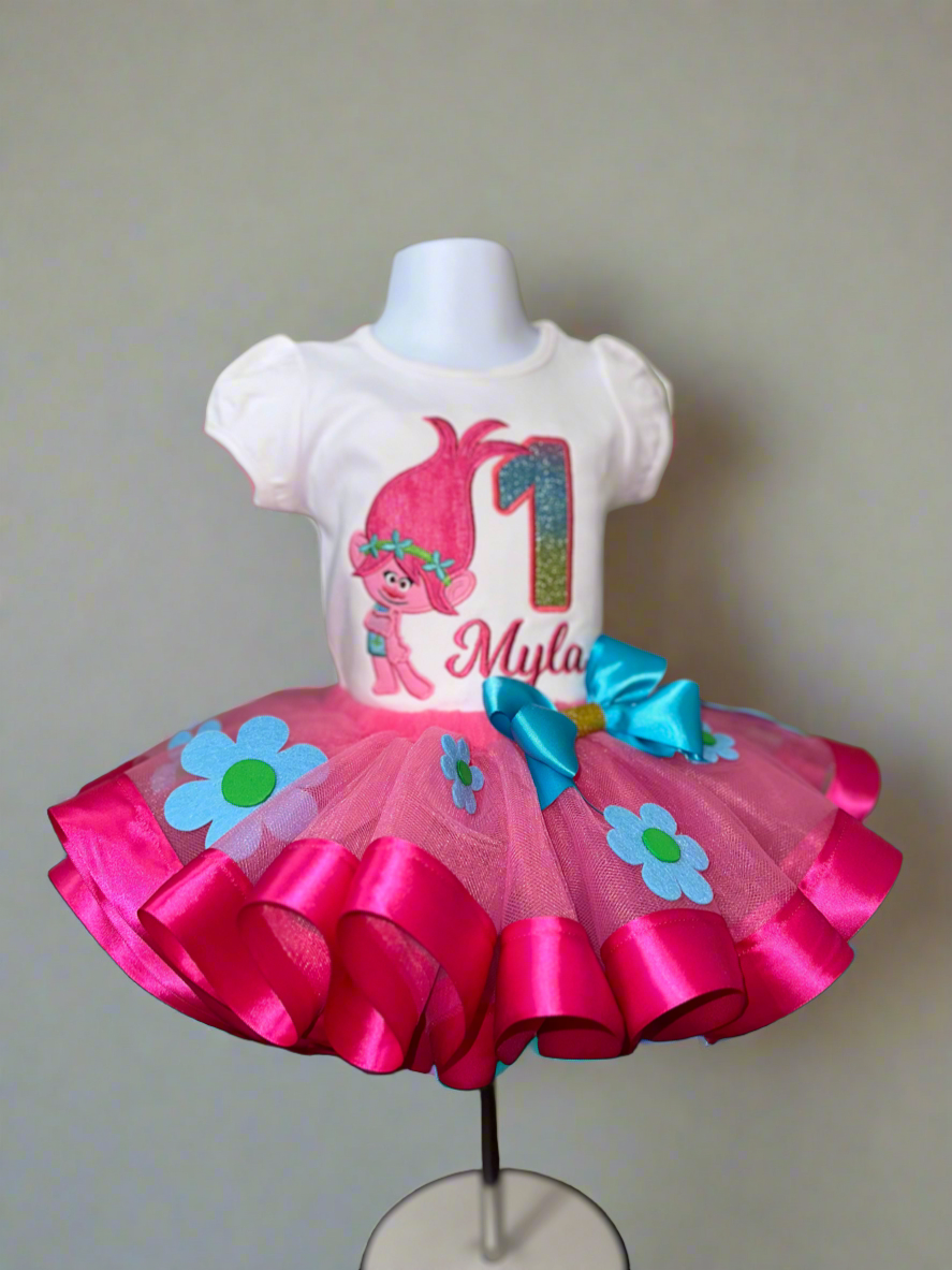 A toddler's birthday outfit featuring a white t-shirt with a pink and green Troll character applique and the name 'Myla' on it, paired with a pink tulle skirt with blue and green flower appliques and a blue bow.