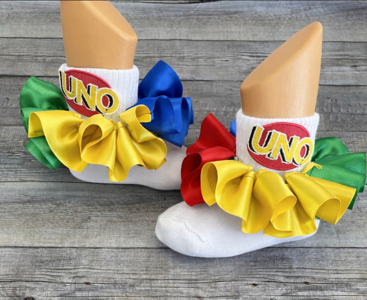 Personalized UNO birthday ruffle socks featuring colorful satin ribbon ruffles and embroidered UNO logos to coordinate with a girl’s UNO birthday outfit.