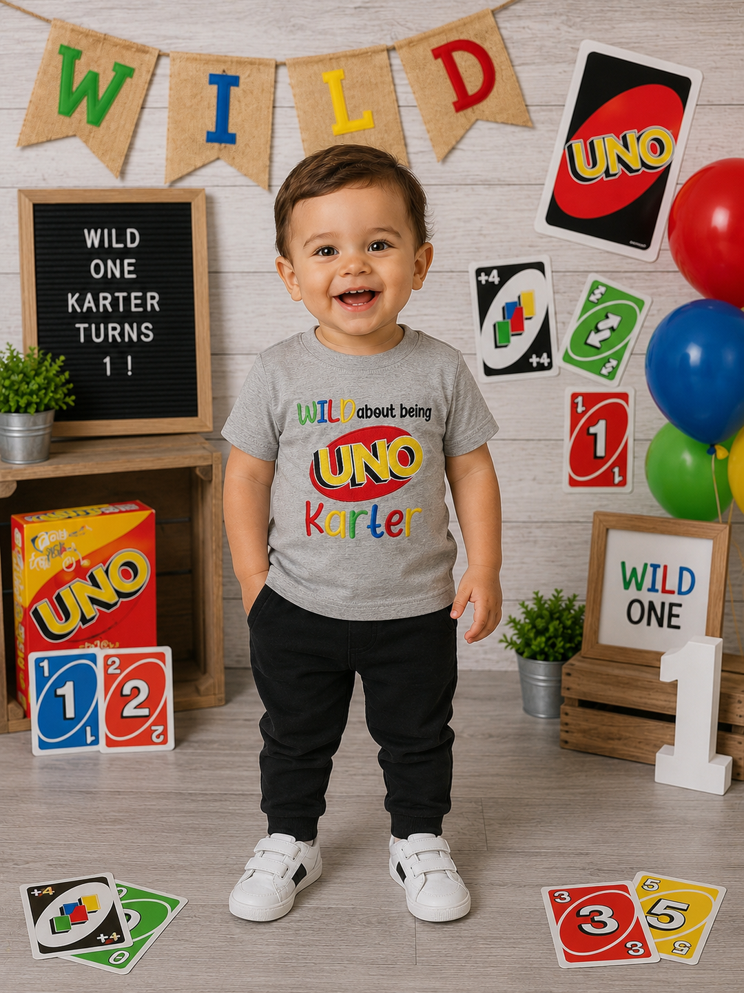 Smiling toddler wearing a gray personalized Wild About Being UNO birthday shirt featuring the UNO logo and custom name at a first birthday party setup.