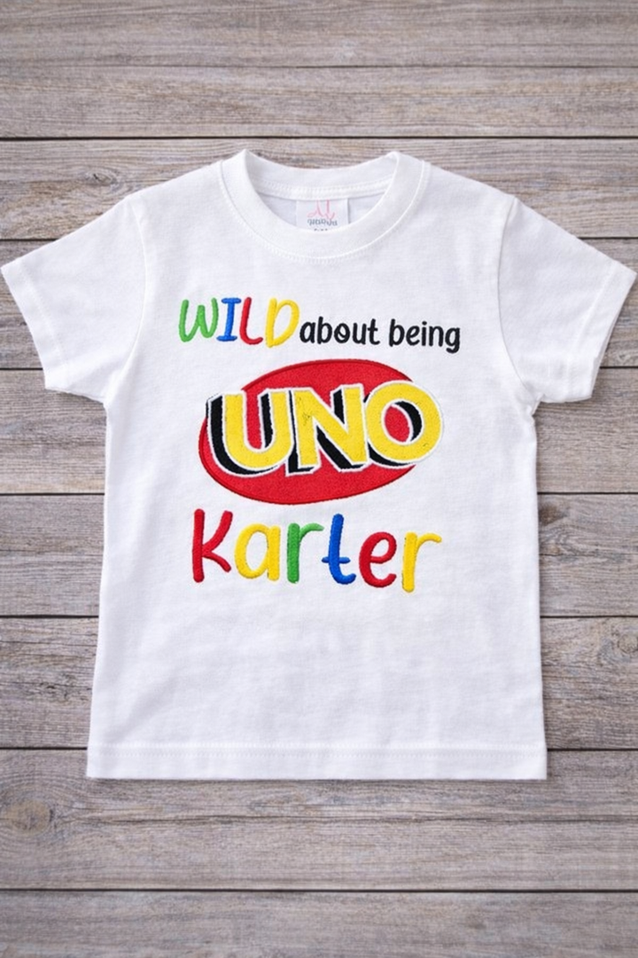 White personalized Wild About Being UNO birthday shirt featuring colorful embroidery, UNO logo design, and custom name for a first birthday outfit.”