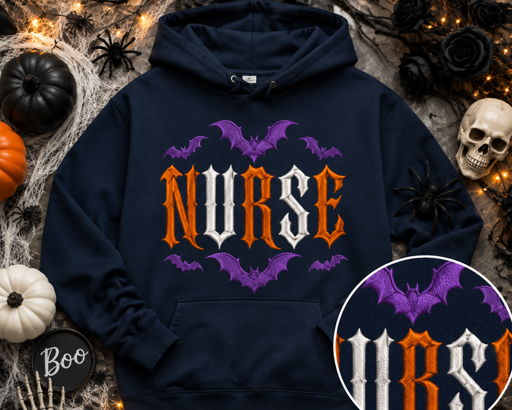 A navy blue hoodie with a Halloween-themed graphic reading 'NURSE' in orange and white gothic lettering, surrounded by purple bats. The hoodie is displayed in a flat-lay arrangement with autumn and Halloween decorations including pumpkins, a skull, spiderwebs, and fairy lights.
